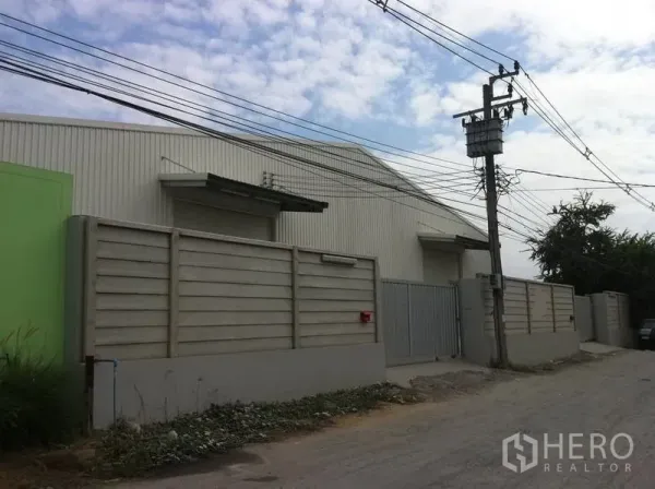 Warehouse for rent in Sai Mai, Bangkok - Street view of gated warehouse compound with high boundary wall, secure metal gate and overhead power lines in Sai Mai, Bangkok.