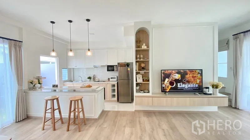 Townhouse for rent in Prawet, Bangkok - Modern white kitchen with island, pendant lights, open shelving and adjacent living area with TV.