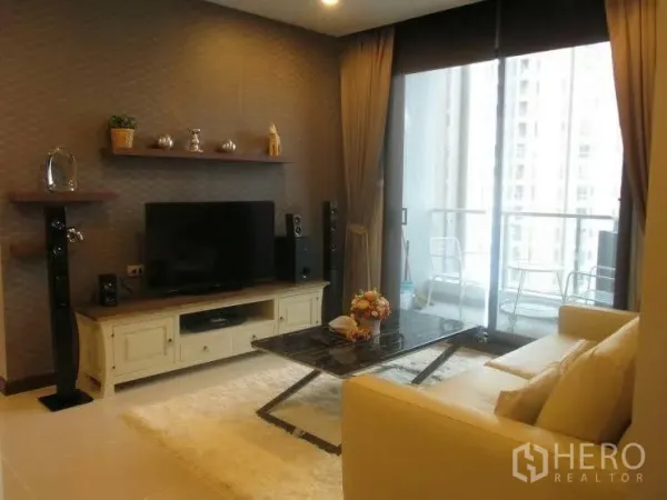 Condominium for rent or sale in Huai Khwang, Bangkok - Contemporary living room with cream sofa, TV console, wall shelves and speakers, opening to a balcony via floor‑to‑ceiling sliding glass doors.