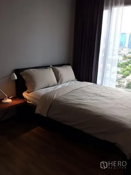Condominium for rent in Watthana, Bangkok - Master bedroom with a double bed, bedside lamp, and floor-to-ceiling window offering a city view.