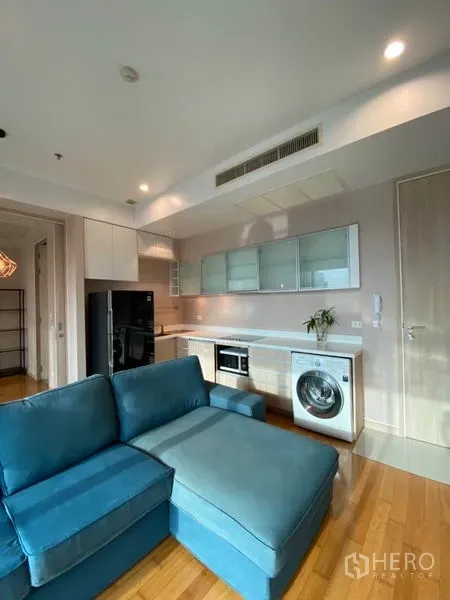 Condominium for rent or sale in Yan Nawa, Bangkok - Compact linear kitchen and L-shaped blue sofa in a bright living room with city outlook.