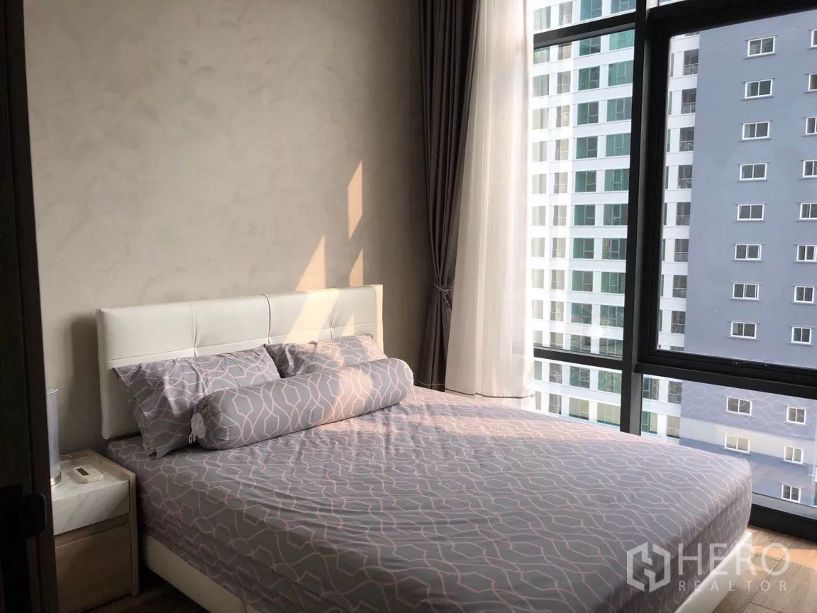 Condominium for sale in Watthana, Bangkok - Second bedroom with a white bedframe, patterned bedding, and corner windows facing a neighboring tower.