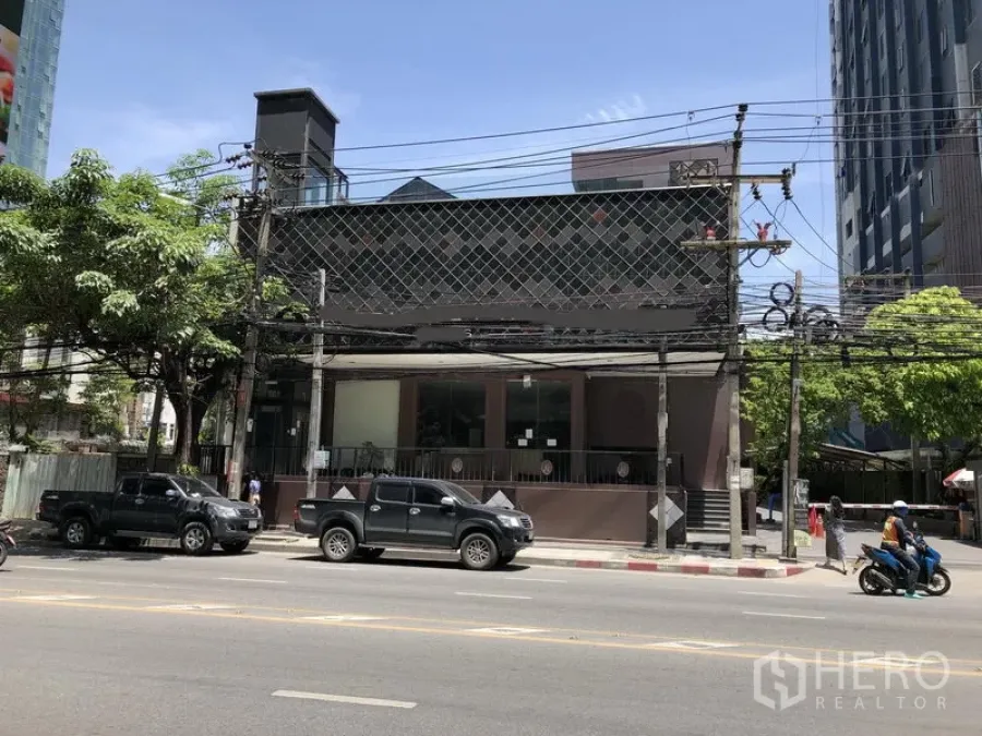 Retail Space for rent in Watthana, Bangkok - Low-rise retail building with wide glass frontage and a raised terrace on Thonglor in Watthana, Bangkok, along a main road with high-rises and trees.