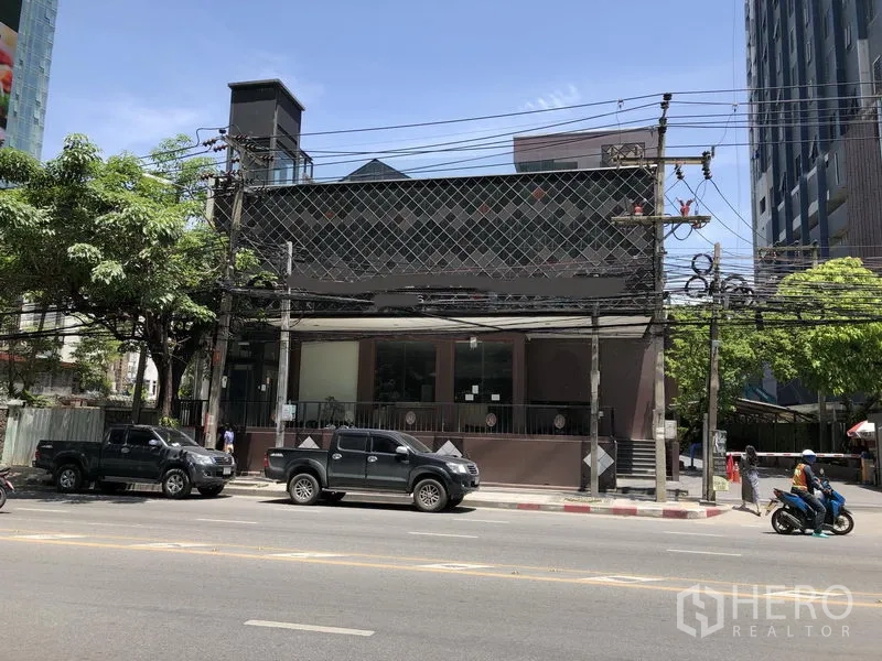 Retail Space for rent in Watthana, Bangkok - Low-rise retail building with wide glass frontage and a raised terrace on Thonglor in Watthana, Bangkok, along a main road with high-rises and trees.