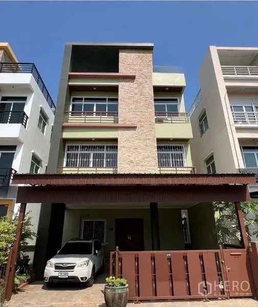 带办公室的家 出租或出售在Min Buri, Bangkok - 三层居家办公楼外立面，砖饰面、阳台与带顶车位及金属大门。