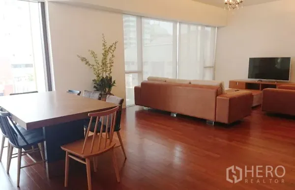 Condominium for sale in Pathum Wan, Bangkok - Open-plan living and dining room with floor-to-ceiling windows, hardwood floors, tan sofas and a TV.