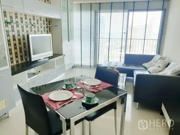 Condominium for rent in Khlong Toei, Bangkok - Bright living and dining area with TV wall, glass table set, leather sofa, and city-view balcony with blinds.