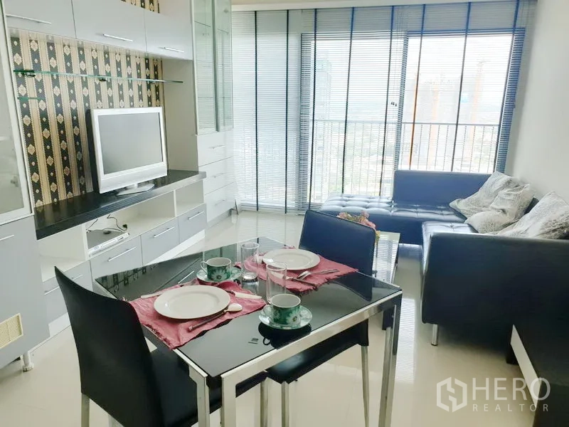 Condominium for rent in Khlong Toei, Bangkok - Bright living and dining area with TV wall, glass table set, leather sofa, and city-view balcony with blinds.