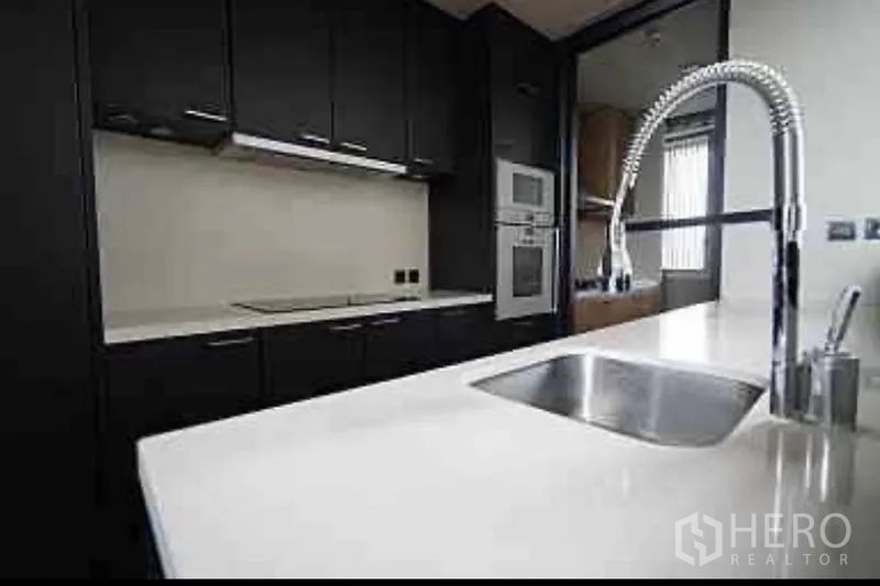 Condominium for rent in Sathon, Bangkok - Kitchen worktop with stainless sink and spring faucet facing dark cabinets and integrated appliances.