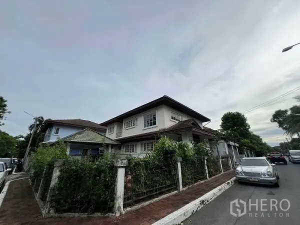 独立式住宅 出售在Phra Khanong, Bangkok - 转角视角，可见围墙与绿色植被以及宽人行道，社区氛围宁静。