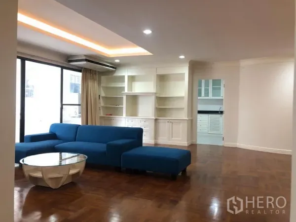 公寓 出租在Watthana, Bangkok - 宽敞客厅，蓝色转角沙发、内置白色书柜、木地板与大落地推窗