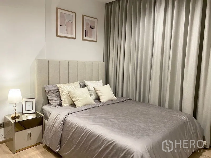 Condominium for rent or sale in Watthana, Bangkok - Bedroom with padded headboard, full drapes, bedside table lamp and neutral decor.