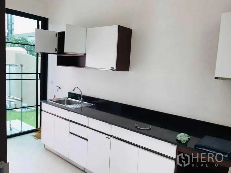 Detached House for rent or sale in Thanyaburi, Pathum Thani - Light kitchen with black stone countertop, white cabinets and a glass door to the backyard.