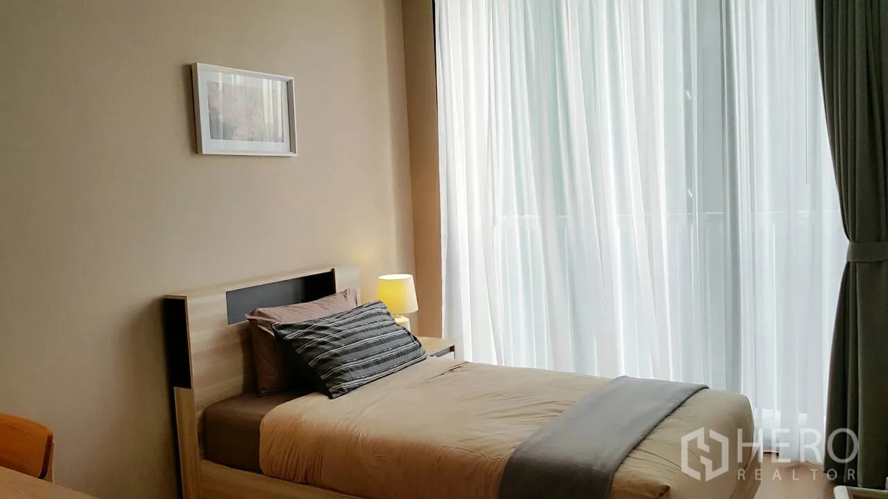 Condominium for rent or sale in Khlong Toei, Bangkok - Second bedroom with a single bed beside sheer floor-to-ceiling curtains and warm lighting.