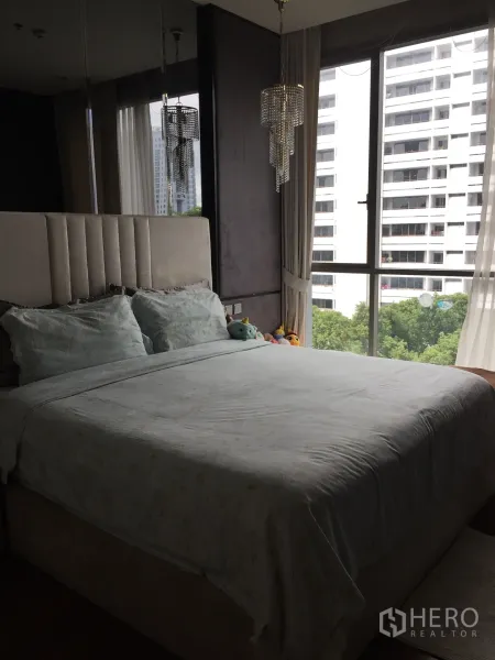 คอนโด ขายในWatthana, Bangkok - ห้องนอนใหญ่มีหัวเตียงบุผ้า โคมระย้า และหน้าต่างมุมกว้างมองเห็นต้นไม้และตึกสูง