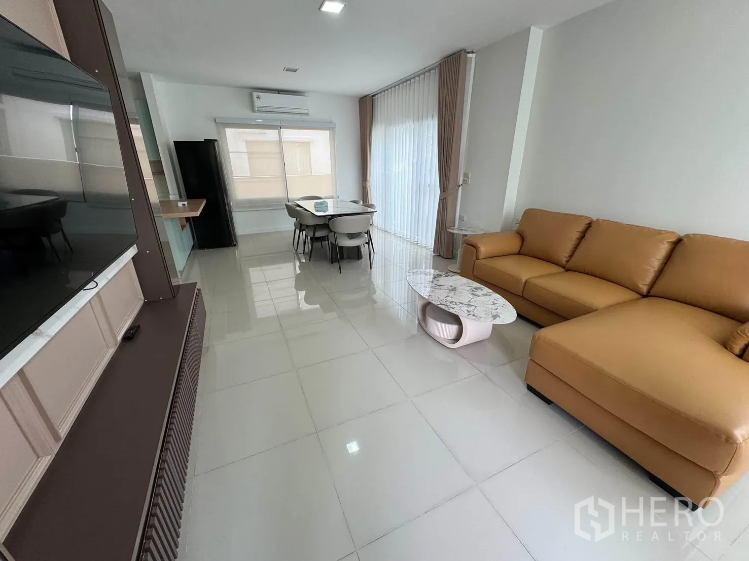 Detached House for rent in Prawet, Bangkok - Living room view with a tan sectional sofa, TV feature wall, and glossy tiled floors.