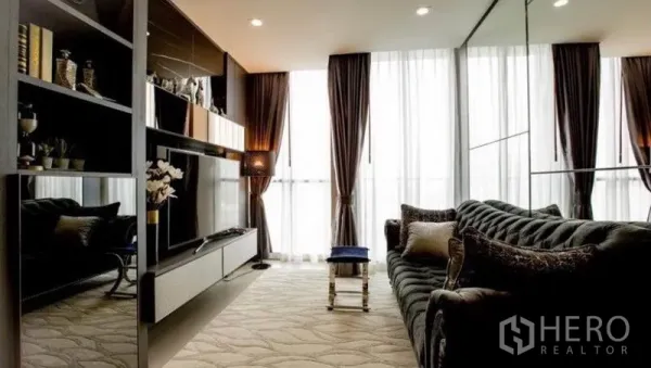 Condominium for rent in Pathum Wan, Bangkok - Cozy living room with dark velvet sofa, shelving and large windows with sheer curtains.