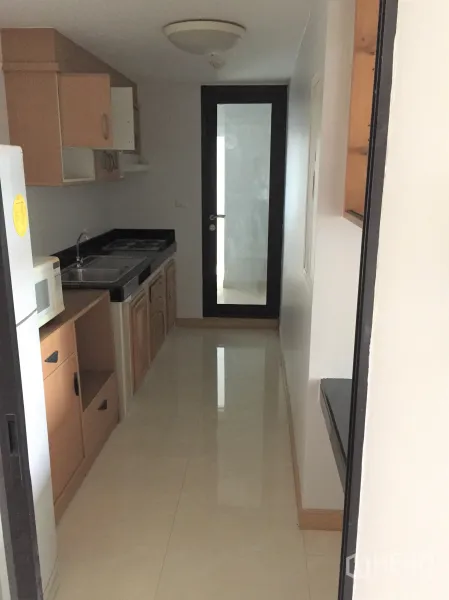公寓 出租在Watthana, Bangkok - 独立走廊式厨房，木质橱柜、微波炉、水槽与黑色台面，玻璃门通往阳台。