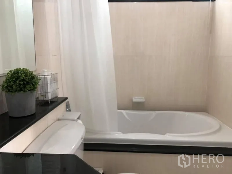 Condominium for rent in Watthana, Bangkok - Modern bathroom with bathtub, white shower curtain and a black countertop with a potted plant.