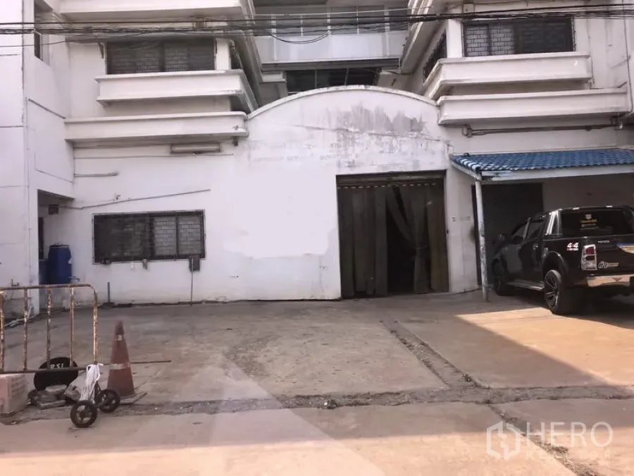 Factory for sale in Lam Luk Ka, Pathum Thani - White factory exterior in Lam Luk Ka with concrete loading yard, wide roll-up door, and blue awning parking beside a black pickup.