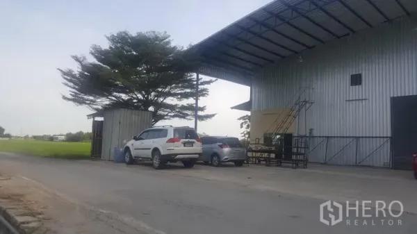 Factory for rent in Khlong Luang, Pathum Thani - Roadside view of the warehouse entrance showing covered loading bay, parked cars, and a tree-lined open field.