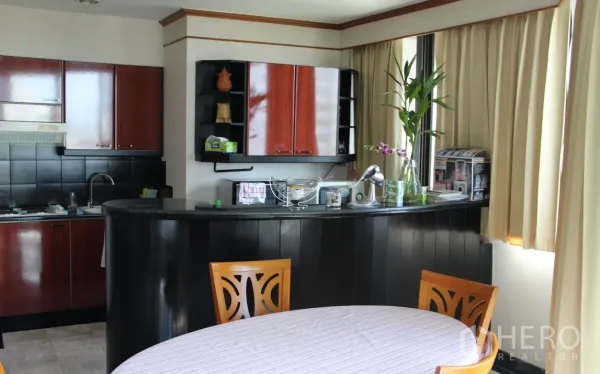Condominium for rent in Khlong Toei, Bangkok - Open kitchen with dark cabinetry, black tile backsplash, and a curved breakfast bar beside a windowed dining area.
