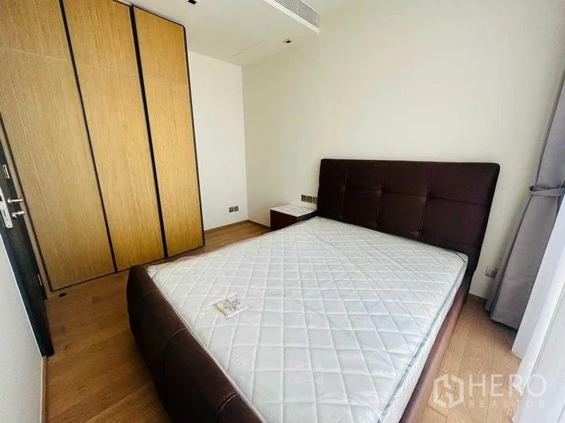 Condominium for rent in Khlong Toei, Bangkok - Cozy bedroom with dark leather bedframe, wood wardrobe, and natural light.