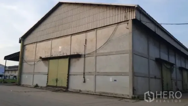 Warehouse for rent in Mueang Pathum Thani, Pathum Thani - Exterior of a large concrete warehouse with a gable roof and green sliding doors in Pathum Thani.