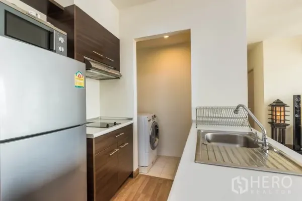 Condominium for rent in Bang Na, Bangkok - Compact kitchen with stainless refrigerator, cooktop, sink, and a laundry nook with washing machine.