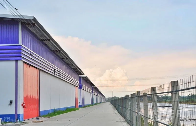 Warehouse for rent in Pathum Thani - Side perspective of warehouses with red doors along a secure perimeter fence beside a service road and waterway.