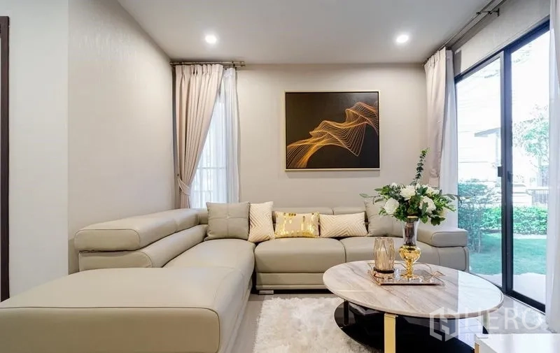 Detached House for sale in Lam Luk Ka, Pathum Thani - Cozy lounge with sectional sofa, artwork and garden view through glass doors.