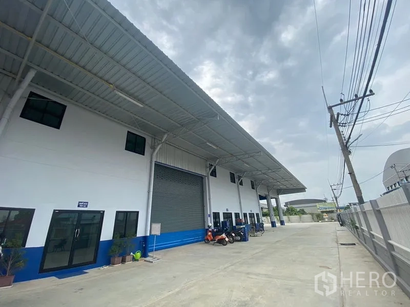 Warehouse for rent in Pathum Thani - Frontage with roller shutter, glass office door, and wide apron under the canopy.