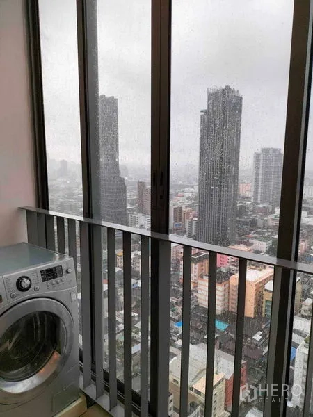 Condominium for rent in Ratchathewi, Bangkok - Enclosed balcony laundry area with washing machine and skyline view through tall windows.