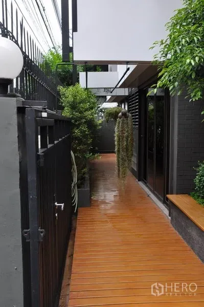 Detached House for rent in Watthana, Bangkok - Wooden deck side walkway bordered by greenery and a black metal fence.