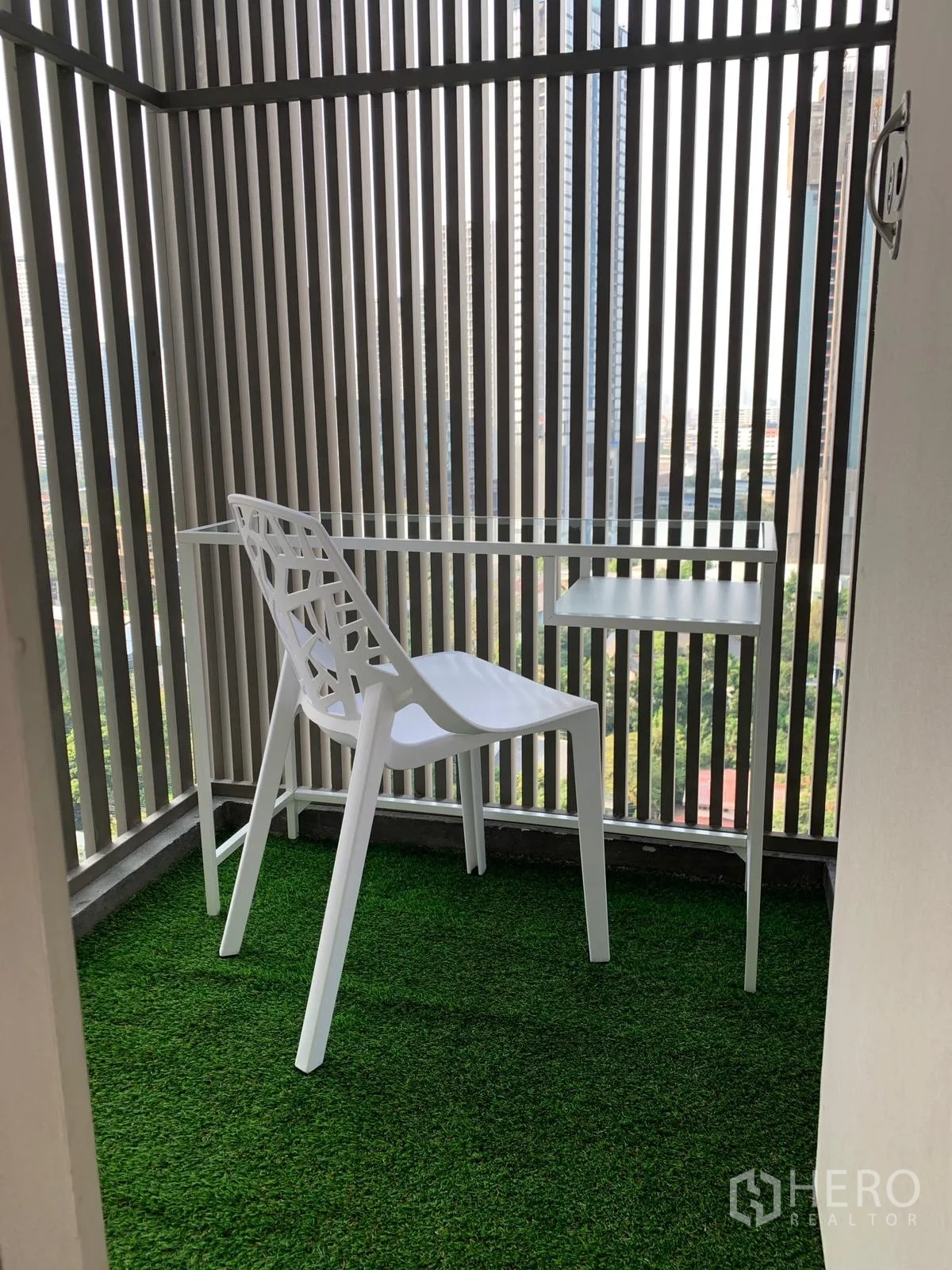 Condominium for sale in Khlong Toei, Bangkok - Compact balcony with artificial grass, white chair and table with a city backdrop.
