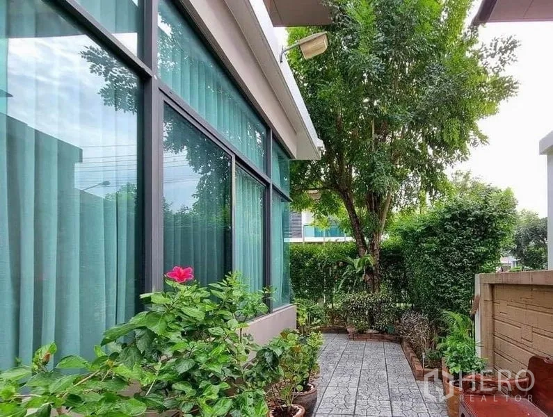 Detached House for sale in Prawet, Bangkok - Side garden path with potted plants beside large curtain windows.