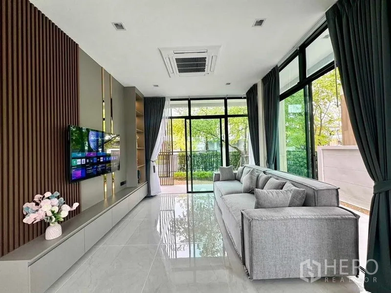 Detached House for rent or sale in Bang Phli, Samut Prakan - Bright living room with large glass doors, ceiling cassette air conditioner, wall-mounted TV and gray sofa facing the garden.