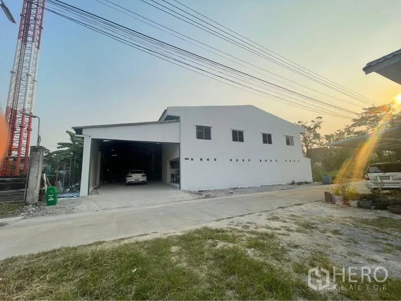 Warehouse for sale in Lam Luk Ka, Pathum Thani - Street view of modern white warehouse with wide access road and off‑street parking at sunset.
