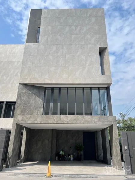 带办公室的家 出售在Prawet, Bangkok - 现代三层家居办公楼立面，灰色石材外墙与有顶车位，蓝天下外观。