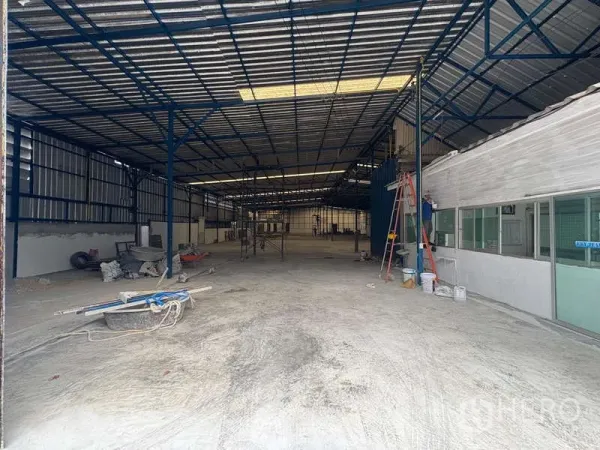Warehouse for rent in Prawet, Bangkok - Entrance view of the warehouse showing concrete flooring, corrugated metal walls, a ladder, and a glass-partitioned office block.