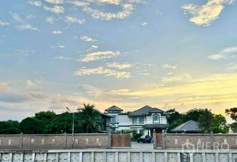 Detached House for sale in Khlong Luang, Pathum Thani - Wide compound view of the residence and boundary wall seen across a canal at sunset.