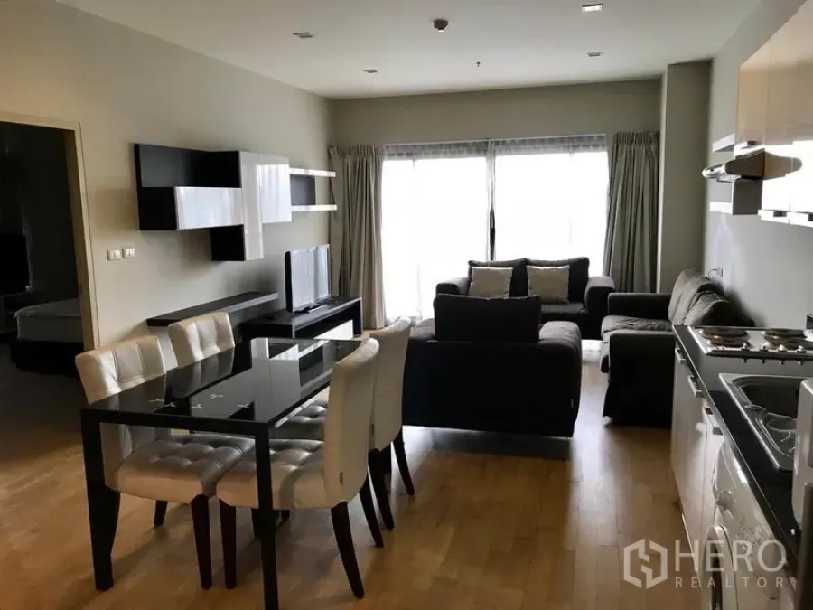Condominium for rent or sale in Watthana, Bangkok - Open-plan living and dining area in a Watthana condo with a glass table, cushioned chairs, dark sofas, TV unit, wall shelves, and large sliding windows with curtains.