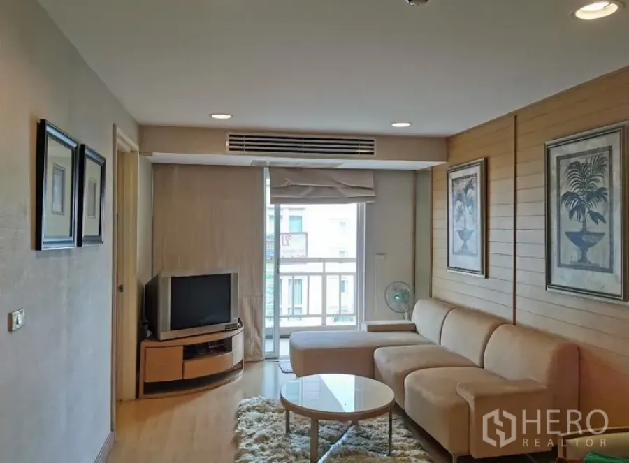 Condominium for sale in Sathon, Bangkok - Bright living room with beige L-shaped sofa, TV console, wall art and sliding door to a small balcony in a Sathon condo.