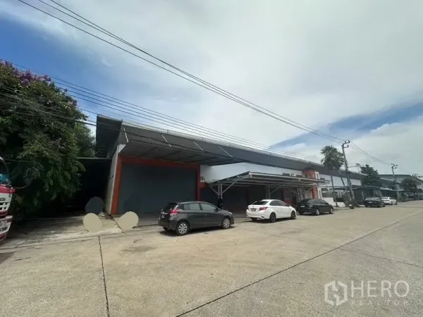 Factory for rent in Nonthaburi - Wide street view of a low-rise factory with roller shutter and parking bays under a steel canopy.