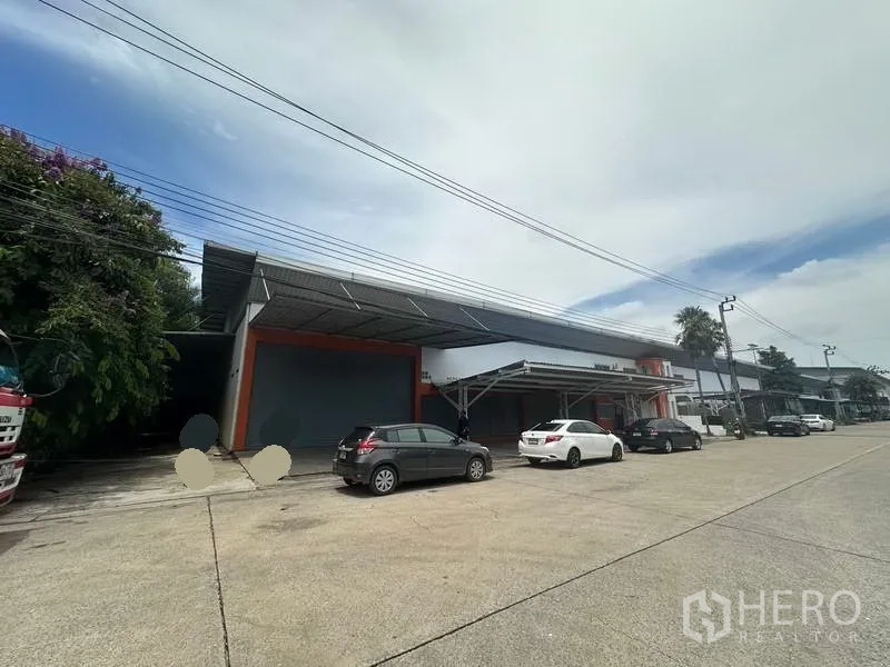 Factory for rent in Nonthaburi - Wide street view of a low-rise factory with roller shutter and parking bays under a steel canopy.
