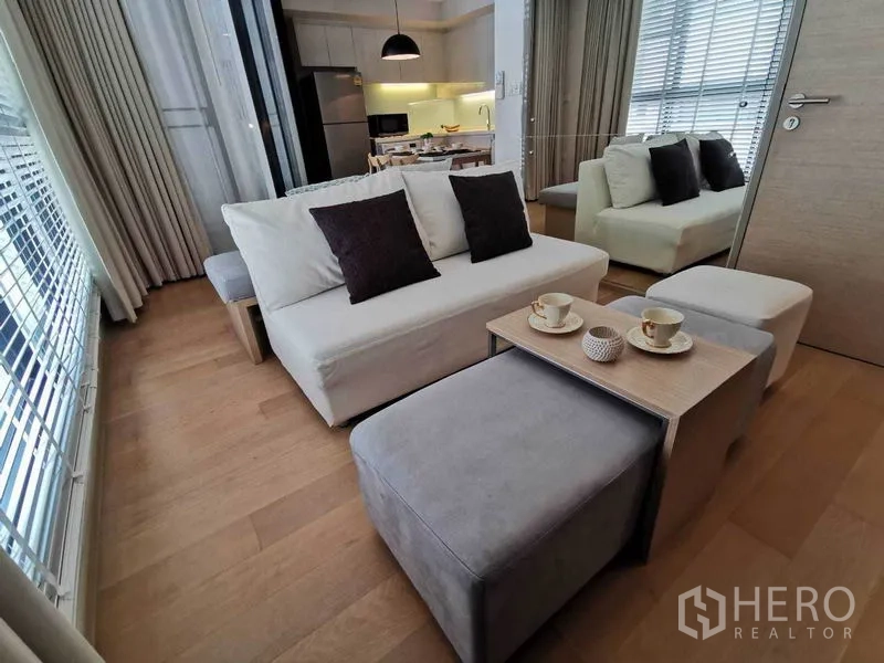 คอนโด ให้เช่าในWatthana, Bangkok - ห้องนั่งเล่นสว่างด้วยโซฟาสีขาว ออตโตมัน ผนังกระจก และพื้นไม้ ข้างครัวเปิดโล่ง