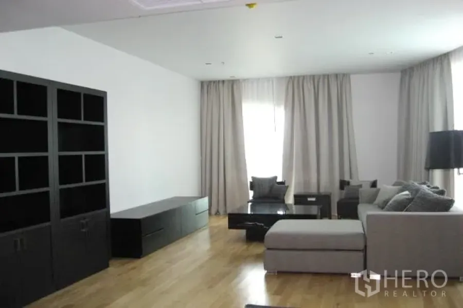 Condominium for sale in Khlong Toei, Bangkok - Spacious living room with grey sectional sofa, black media console and shelving, light wood floors, and large curtained windows.