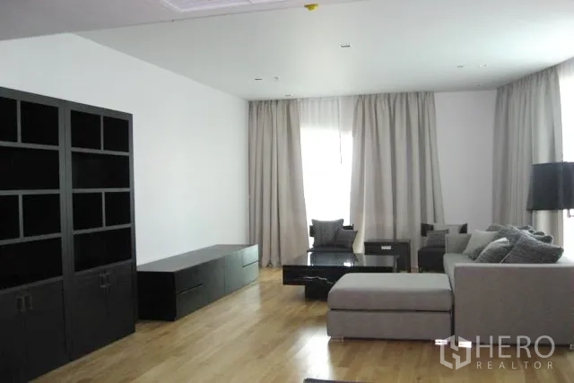 Condominium for sale in Khlong Toei, Bangkok - Spacious living room with grey sectional sofa, black media console and shelving, light wood floors, and large curtained windows.