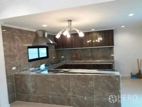 Detached House for rent or sale in Prawet, Bangkok - U-shaped marble-look kitchen with range hood, overhead cabinets and chandelier lighting.