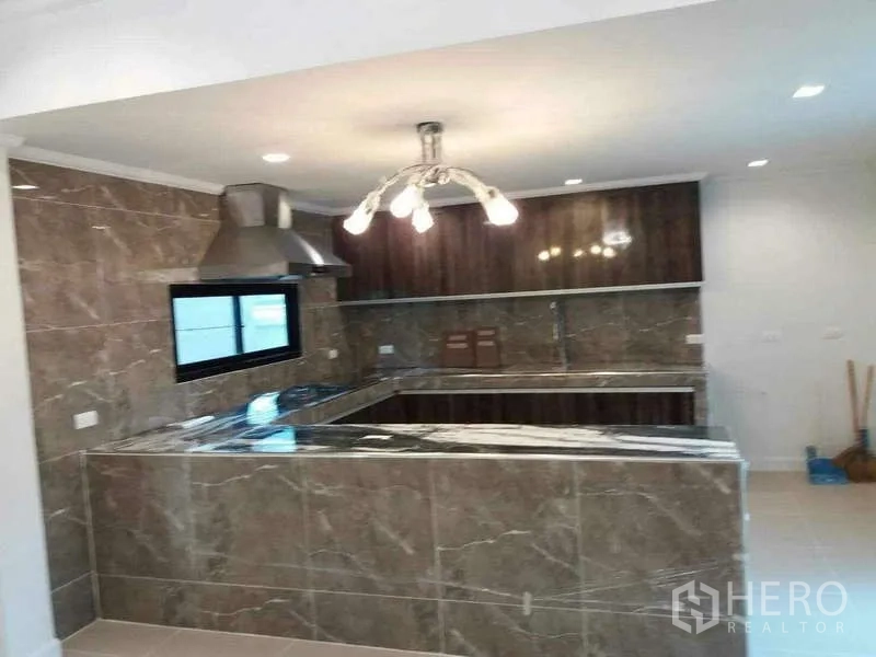 Detached House for rent or sale in Prawet, Bangkok - U-shaped marble-look kitchen with range hood, overhead cabinets and chandelier lighting.